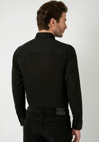 Zwarte langemouwen blouse met een subtiel geometrisch patroon, voorzien van een puntige kraag en knoopmanchetten, gecombineerd met donkere jeans.