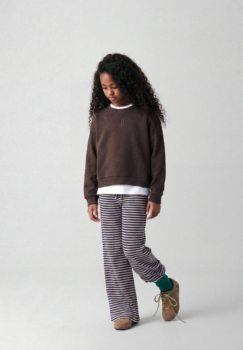 Jeune fille aux cheveux bouclés porte un sweat-shirt marron foncé, un pantalon rayé, des chaussettes vertes et des chaussures marron, debout, la tête légèrement baissée.
