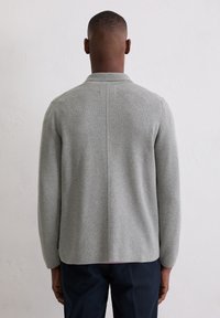 Ein Mann trägt einen hellgrauen Langarmpullover und dunkle Hosen und steht vor einer schlichten weißen Texturwand.