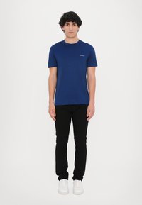 Tamsiai mėlynas medvilninis marškinėliai su apykakle ir trumpomis rankovėmis, derinamas su juodomis siauriais kelnėmis ir baltomis sportinėmis bateliais, be matomų raštų.