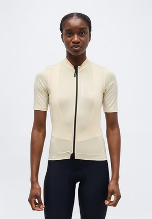 Γυναίκα ντυμένη με μπεζ ποδηλατική φανέλα με κοντό μανίκι και φερμουάρ στο μπροστινό μέρος, και μαύρο ποδηλατικό σορτς, στέκεται μπροστά σε λευκό απλό φόντο.