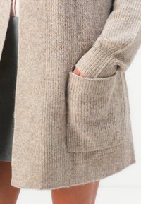 Hand in der Tasche eines beigen Strickcardigans mit gerippten Bündchen und strukturiertem Stoff über nackten Beinen.
