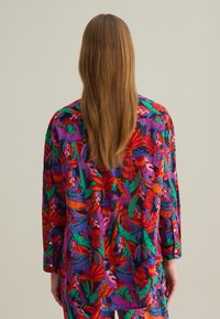 Blusa floral multicolorida con un corte holgado, que presenta patrones vibrantes en rojo, verde y púrpura. Mangas largas y detalles de botones.