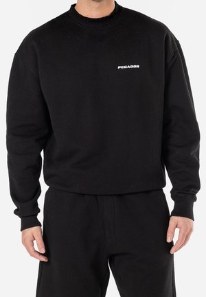 Hombre con sudadera negra de manga larga con el logo "PEGADOR" en el pecho y pantalones negros a juego.