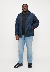 Marineblå vindjakke med hette, elastiske mansjetter, frontlukking med glidelås, kombinert med en svart t-skjorte og lyseblå jeans, samt svarte joggesko.
