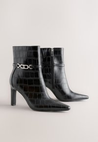 Black crocodile-patterned leather ankle boots with pointed toes, mid-height heels, side zipper, and silver chain detail on neutral background.