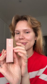 ILIA Skin Tint, en slank lyserød æske med hvid tekst, holdt i en hånd. Materialet virker glat med et minimalistisk design, der viser produktinformation.