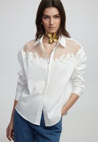 Weißes Hemd mit Knopfleiste und Netz-Einsatz mit floraler Stickerei. Kombiniert mit Jeanshose und einem aufwendigen goldenen Schmetterlings-Halsband.