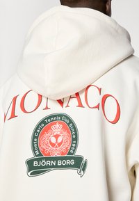 Biela mikina s kapucňou s grafikou v zelenej a červenej farbe, ktorá zobrazuje "MONACO" a "BJÖRN BORG" spolu s korunovaným emblémom Monte Carlo Tennis Club.