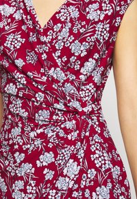 Burgundy omlottklänning med vita blommönster, korta ärmar och knytdetalj i midjan. Tyget verkar mjukt och strukturerat.