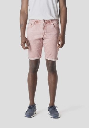 Mies, joka pukeutunut vaaleanpunaiseen käännettyihin farkkushortseihin, valkoiseen t-paitaan ja tummansinisiin tennareihin, seisoo yksinkertaista vaaleaa taustaa vasten.