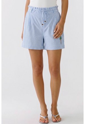 Frau trägt hellblaue und weiße gestreifte Shorts mit gebundenem Bund und Palmen-Stickerei, kombiniert mit silbernen Sandalen und einem weißen Oberteil.