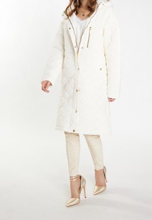 Femme portant un long manteau matelassé blanc avec des boutons et fermetures éclair dorés, un pantalon beige et des chaussures à talons hauts dorés, debout sur un fond blanc.