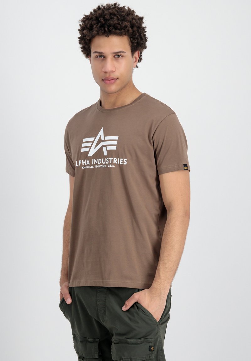 Hnedé tričko s krátkym rukávom s bielou potlačou loga "ALPHA INDUSTRIES." Vyrobené z jemnej bavlny, má klasický strih s okrúhlym výstrihom.