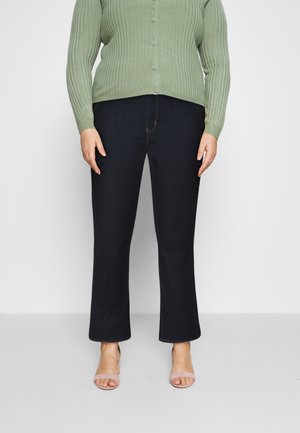 Donkerblauwe denim jeans met een rechte pijp, gecombineerd met een lichtgroene geribde cardigan en beige sandalen. Knopen zijn zichtbaar op de cardigan.