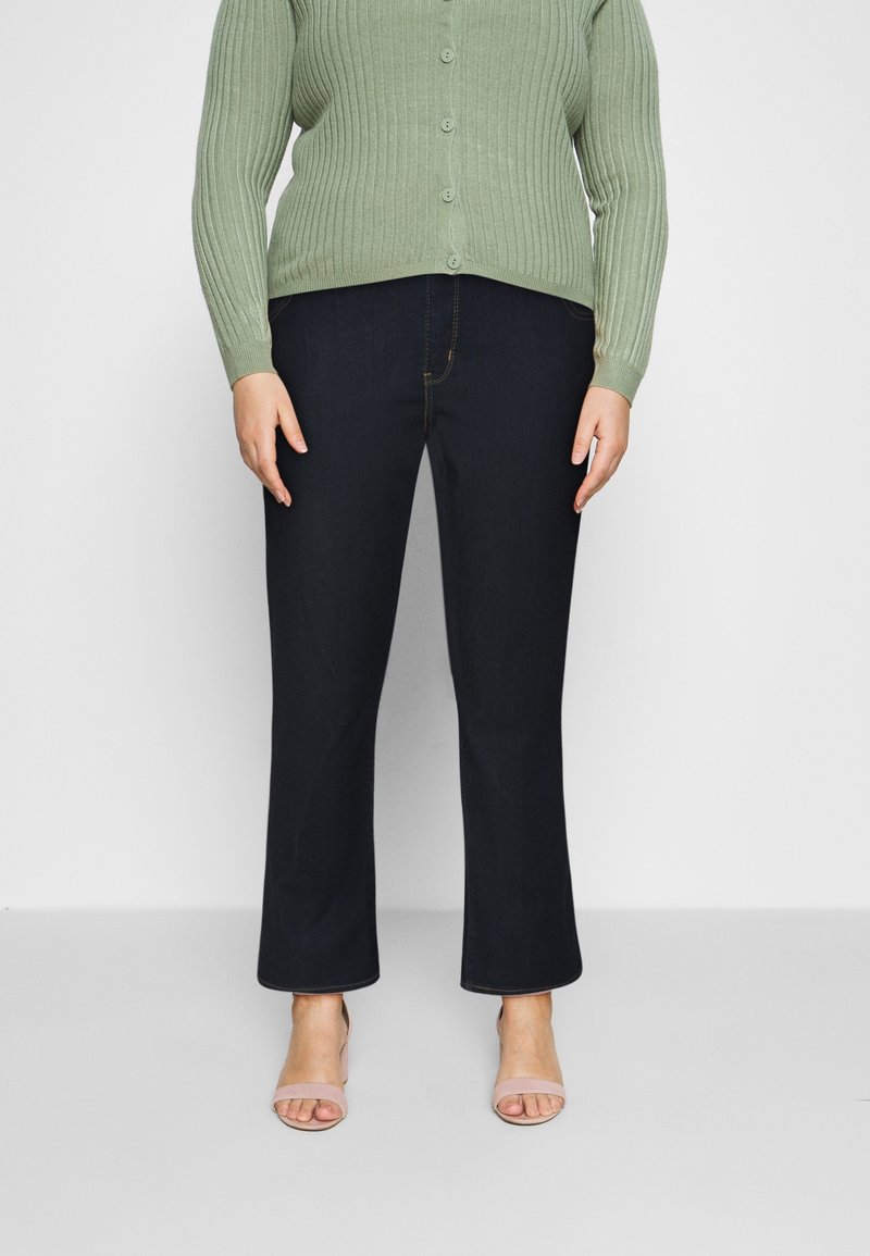 Vaqueros de mezclilla azul oscuro con pierna recta, combinados con un cárdigan de canalé verde claro y sandalias beige. Botones visibles en el cárdigan.