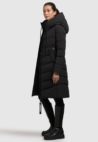 Abrigo de invierno acolchado negro con un cuello alto, cierre de cremallera, bolsillos laterales y un diseño de acolchado texturizado. Lleva puesto con botas negras hasta la rodilla.