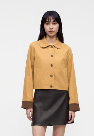 Vrouw met steil zwart haar draagt een mosterdgele jas met knopen en bruine manchetten en een zwarte leren rok, staand tegen een effen achtergrond.