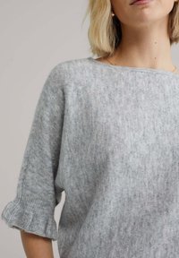 Pull tricoté gris clair avec une texture douce, présentant une coupe décontractée et des manches à volants. L'encolure est large et légèrement de style ras du cou.