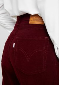 Pantalon en velours côtelé bordeaux avec une étiquette de marque en cuir, doté d'un design de poche arrière signature et de surpiqûres contrastées.