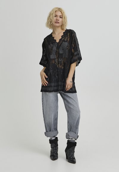 Top ample en dentelle noire avec col en V et manches courtes, présentant un motif sportif et un design superposé. Associé à un jean clair retroussé et des bottines noires.