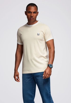 Helles beige T-Shirt mit kurzen Ärmeln, weißen Akzenten und kleinem Logo auf der Brust, kombiniert mit blauen Jeans und einer schwarzen Uhr.