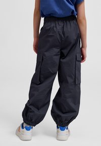 Copil purtând pantaloni cargo negri cu manșete elastice și adidași albi cu accente albastre, stând cu spatele, pe un fundal simplu.