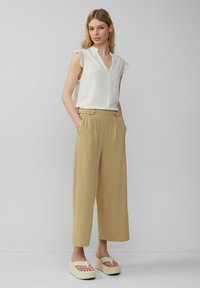 Witte blouse met kantdetails, beige wijde broek met zijzakken en knoopdetails, en beige platform sandalen. Eenvoudige achtergrond.