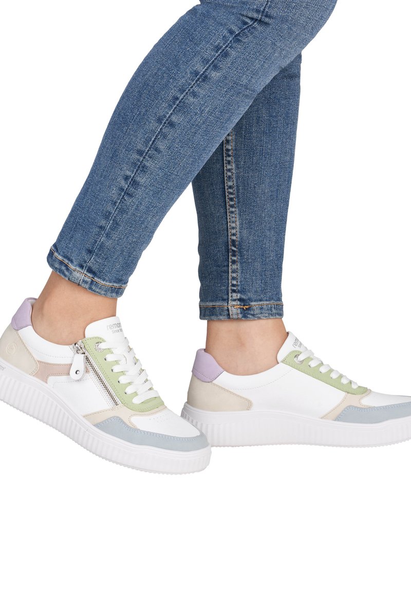 Personne portant un jean bleu et des baskets blanches avec des accents violets, verts, beiges et gris, dotées d'une fermeture éclair latérale sur une chaussure.