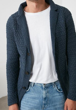 Mann trägt einen strukturierten marineblauen Strickblazer über einem schlichten weißen T-Shirt und hellblauen Jeans, steht drinnen.
