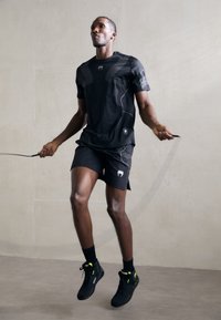 Sort atletisk skjorte med mesh-detaljer, parret med sorte shorts. Modellen hopper med en sjippetov, iført sorte sko med neonlys.
