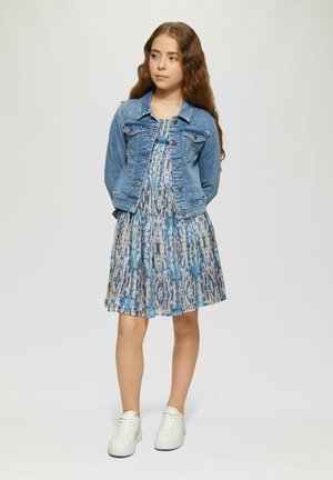 Jeansjacke über einem blau-grün gemusterten Kleid mit Plissee-Design. Das Model trägt weiße Sneakers.