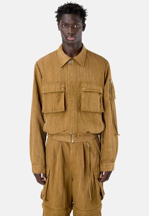 Jonge man die een beige werkjack draagt met grote klepzakken en bijpassende cargobroek tegen een effen achtergrond.