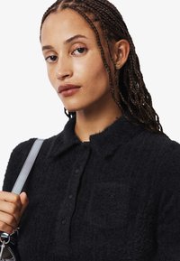 Polo negro texturizado con cuello y botones, que presenta un logo sutil en el bolsillo. El modelo sostiene un bolso con correa plateada.