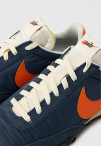 Blaue und orange Nike-Sneaker verfügen über ein Obermaterial aus Nylon, strukturierter Stoff, cremefarbene Schnürsenkel und das auffällige orangefarbene Swoosh-Logo an der Seite.