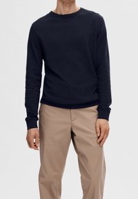 Sweat-shirt bleu marine à manches longues avec col rond, poignets côtelés et coupe décontractée, associé à un pantalon beige clair. Tissu lisse et texturé.