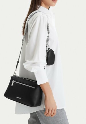 Žena v bílé košili a šedých kalhotách nese černou crossbody tašku Lasocki s odděleným malým pouzdrem na vzorovaném popruhu.