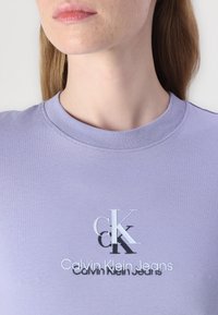 Laventeliväri t-paita, jossa on pyöreä pääntie, lyhyet hihat ja keskellä graafinen kuva, jossa lukee "Calvin Klein Jeans" valkoisella ja mustalla.