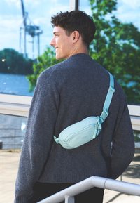 Borsa vita azzurra chiara realizzata in materiale sintetico liscio, decorata con un motivo geometrico, dotata di tracolla regolabile e chiusura a zip, indossata su un maglione grigio.