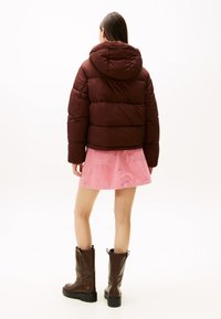 Chaqueta acolchada marrón con capucha, de tejido texturizado y diseño acolchado. Combinada con una falda de pana rosa y botas marrones hasta la rodilla.