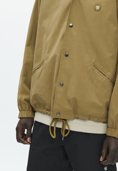 Veste marron boutonnée avec poignets élastiques et ourlet à cordon, portée par-dessus une chemise beige et un pantalon noir, mains détendues le long du corps.