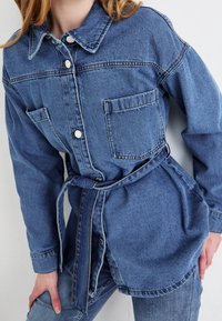 Chaqueta de camisa de denim en azul claro, con cuello, cierre de botones, dos bolsillos en el pecho, cintura con cinturón y mangas largas. Textura suave.