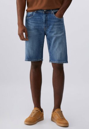 Uomo con pantaloncini di denim blu e sneakers di camoscio color sabbia, in piedi con una mano in tasca contro uno sfondo uniforme.