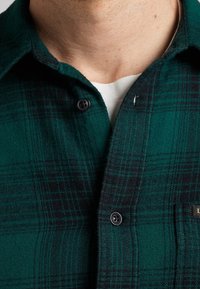 Groene en zwarte geblokte flanellen shirt met een knoopsluiting kraag, voorzien van twee zwarte knopen en een klein logo-tag nabij de zak.