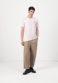 T-shirt rose clair à manches courtes arborant un petit logo jaune sur la poitrine, associé à un pantalon beige ample et des chaussures noires.
