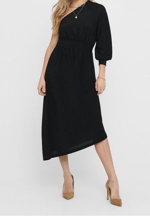 Kobieta ubrana w czarną sukienkę midi na jedno ramię z bufiastym rękawem i brązowe wysokie obcasy, stojąca na jednolitym jasnym tle.