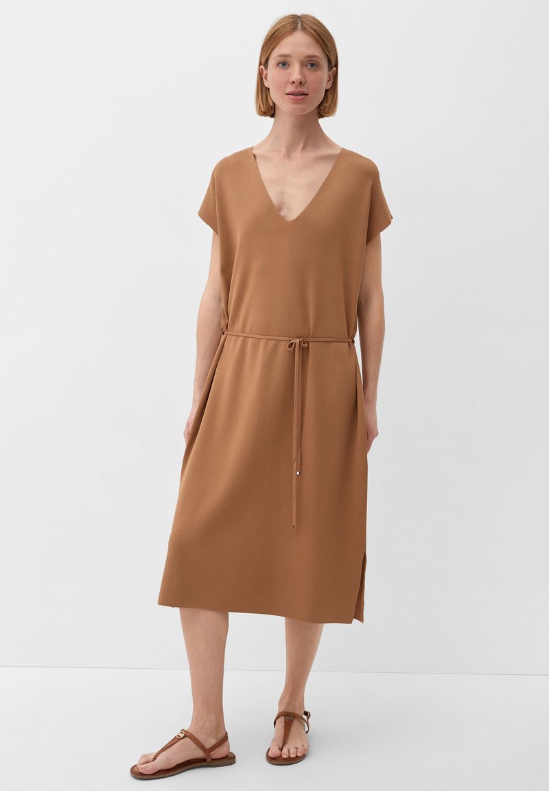 Vrouw in een bruine V-hals midi-jurk met een dunne riem, zijsplit en bruine platte sandalen, staande tegen een effen witte achtergrond.