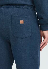 Hombre que lleva pantalones de chándal azul oscuro con cintura elástica y un parche de cuero cuadrado en el bolsillo trasero, mano visible al lado.