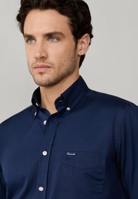 Chemise bleu marine à boutons, avec une petite poche poitrine, des boutons blancs et un accent de logo. Texture lisse et coupe ajustée.