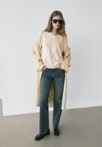 Jonge vrouw in een beige argyle-trui, een lange beige jas, blauwe spijkerbroek, zwarte schoenen en donkere zonnebril staand op een grijze vloer tegen een witte muur.